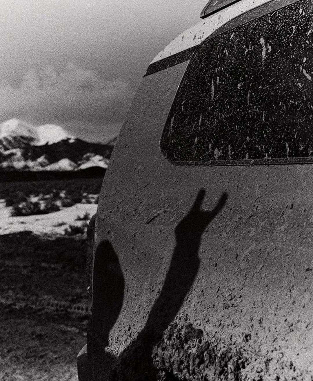 Monochrome image of a dirty SUV's rear with a silhouetted hand making a 'rock on' sign, set against a backdrop of snow-capped mountains.