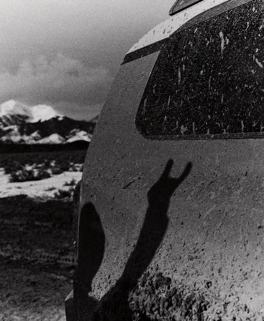 Monochrome image of a dirty SUV's rear with a silhouetted hand making a 'rock on' sign, set against a backdrop of snow-capped mountains.