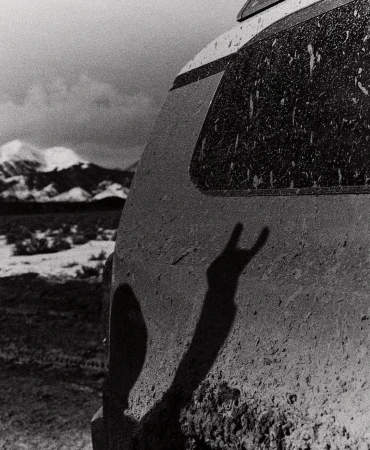 Monochrome image of a dirty SUV's rear with a silhouetted hand making a 'rock on' sign, set against a backdrop of snow-capped mountains.