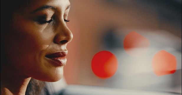 Woman with dark hair and red lipstick in soft, warm lighting, eyes closed, with blurred red and white lights in background. Close-up portrait.