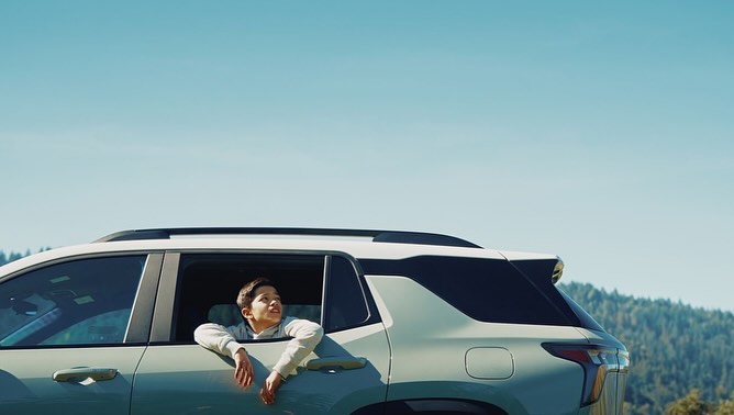 A young boy looking up while leaning out the rear window of a light-colored SUV under a clear blue sky