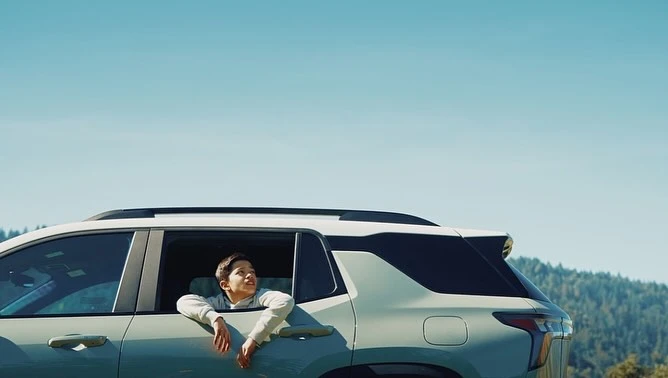 A young boy looking up while leaning out the rear window of a light-colored SUV under a clear blue sky