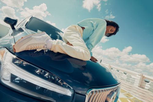 Woman wearing a light blue sweatshirt and cream trousers lying across the hood of a dark blue car against a cloudy sky