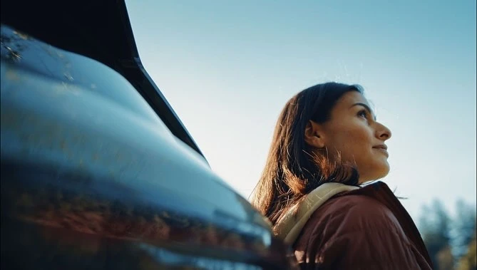 Low-angle profile of a woman leaning against an open car trunk outdoors under a clear blue sky, photographed by Chris Delorenzo.