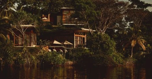 Exterior view of wooden buildings surrounded by lush green trees and vegetation, reflected in the tranquil water of a river.