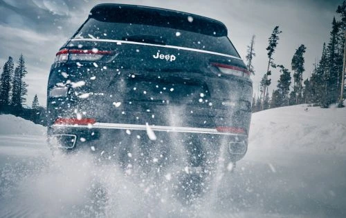 Rear view of a dark blue Jeep SUV driving on snow with trees in the background and snow spraying from the tires.