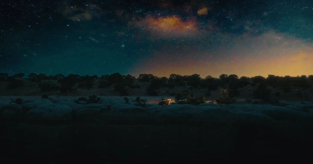 Wide shot captures a camper van with lights on, nestled among trees under a dramatic, starry night sky with a colorful nebula.