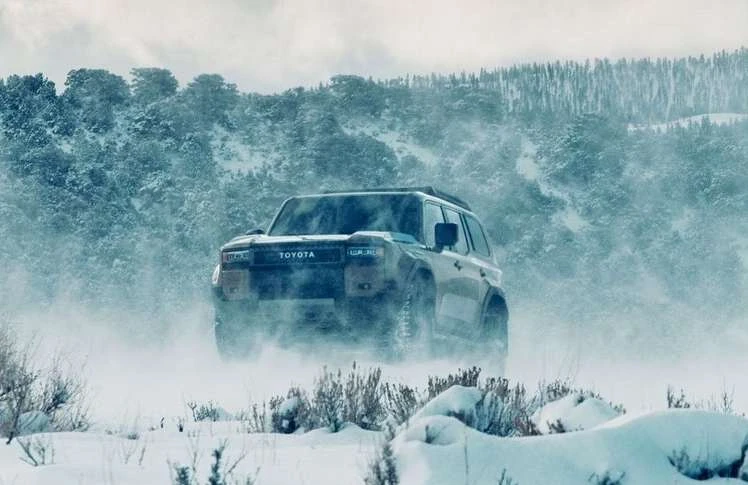 Toyota SUV driving on a snowy landscape with snow-covered mountain and forest in the background, in a winter setting and hazy daylight.