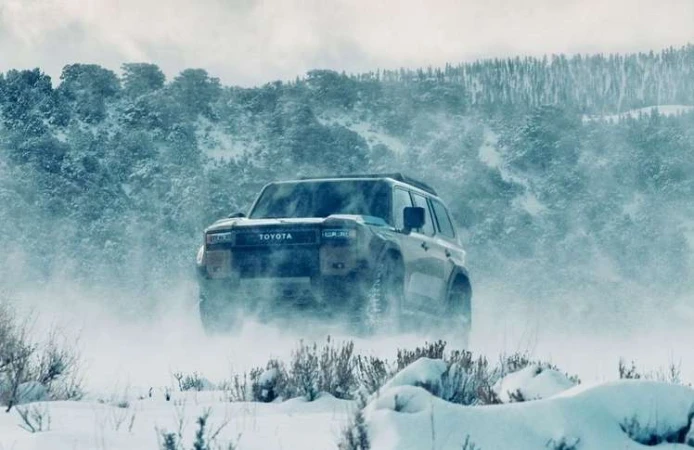 Toyota SUV driving on a snowy landscape with snow-covered mountain and forest in the background, in a winter setting and hazy daylight.