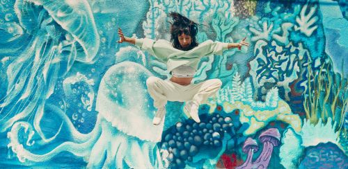 An athletic woman leaps in the air with arms outstretched against a colorful graffiti wall featuring blue jellyfish and sea life.