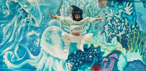 An athletic woman leaps in the air with arms outstretched against a colorful graffiti wall featuring blue jellyfish and sea life.