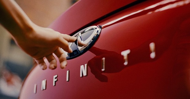 Close-up of a hand touching the chrome logo on the red hood of an Infiniti car, with the car's name spelled out below.