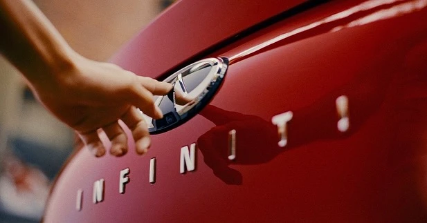 Close-up of a hand touching the chrome logo on the red hood of an Infiniti car, with the car's name spelled out below.
