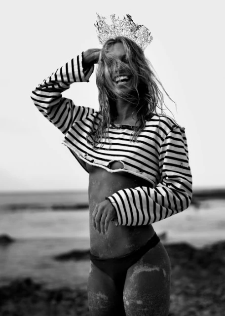 Woman smiles on a beach, wearing a striped crop top, bikini bottoms, and a foil crown, black and white, backlit by the sun.