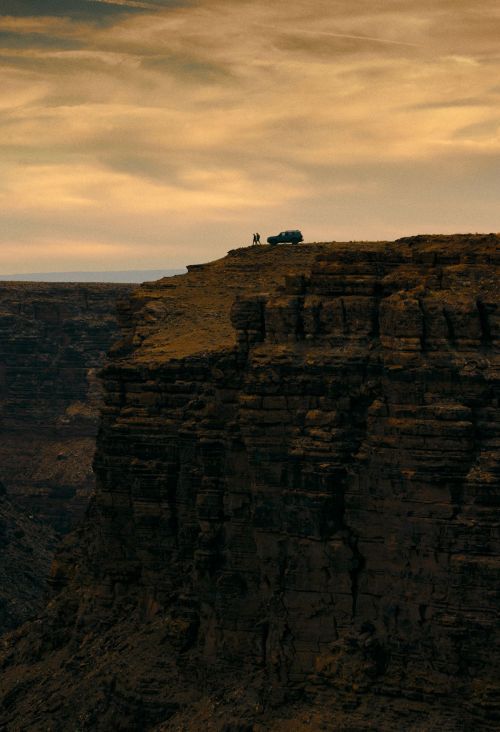 Canyon vista with dramatic cliffs, a vehicle and two people silhouetted on the ridge, under a golden sky.