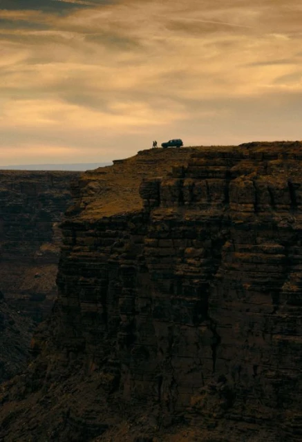 Canyon vista with dramatic cliffs, a vehicle and two people silhouetted on the ridge, under a golden sky.