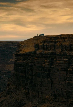 Canyon vista with dramatic cliffs, a vehicle and two people silhouetted on the ridge, under a golden sky.