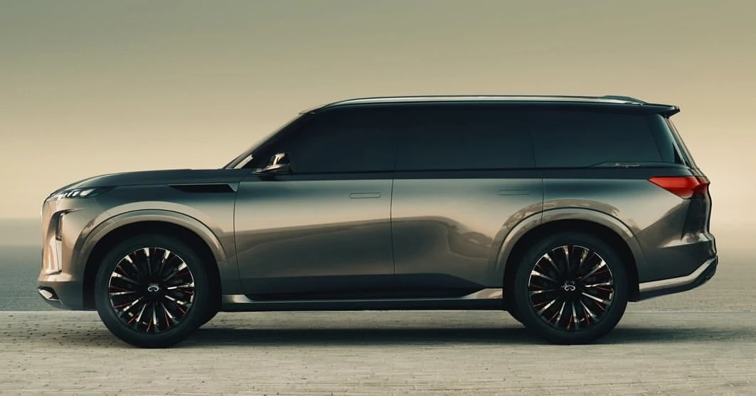 Side view of a dark grey Infiniti SUV on a light grey surface under a bright sky, showcasing its sleek design and large wheels.