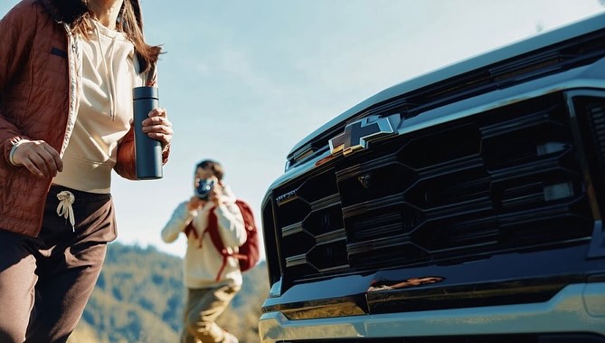 Chevrolet truck showcased outdoors with woman holding a water bottle and a man taking a photo in the background on a sunny day.