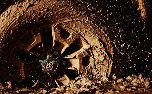 Close-up of a vehicle tire stuck in thick mud, with the Toyota logo visible on the wheel hub. The tire and mud are brown.
