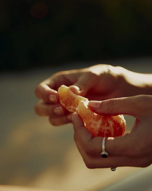 Close-up of hands peeling a juicy mandarin orange segment outside in warm light, highlighting its vibrant color and freshness.
