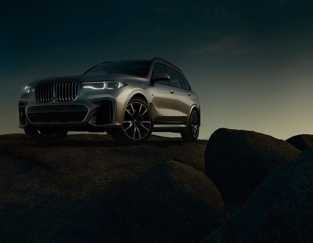Silver BMW X7 SUV parked on a rocky surface under a dim sky, showcasing luxury automotive design and outdoor setting.