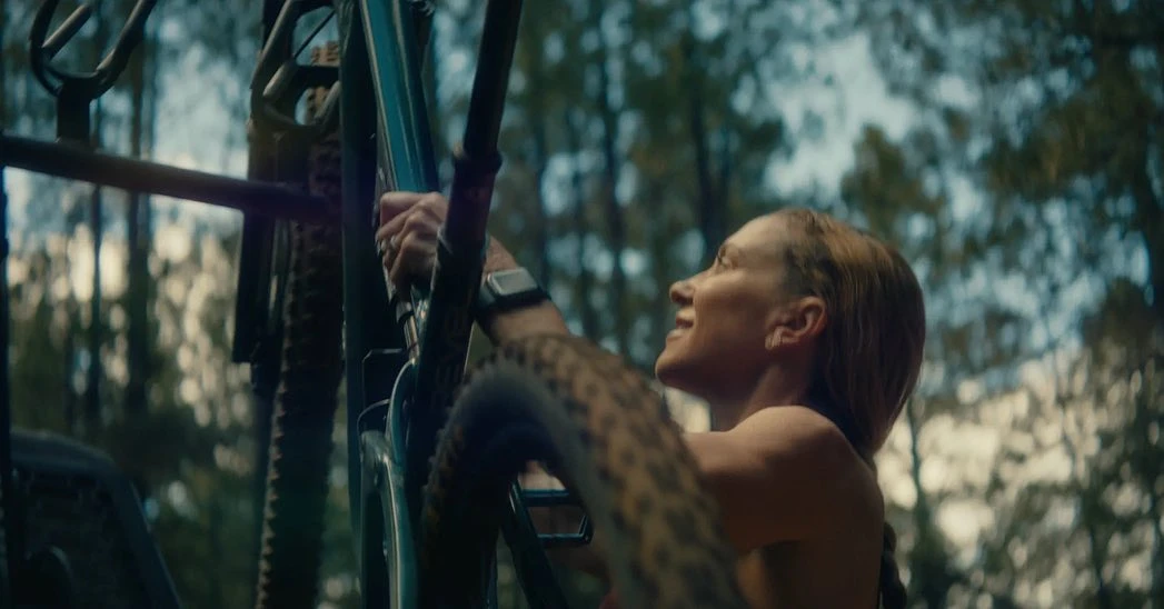 Woman smiling outdoors with bicycle in a forest setting with dappled sunlight filtering through the trees, creating a natural, sporty scene.
