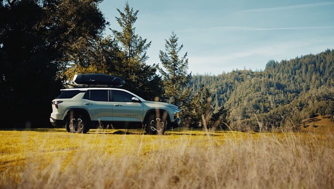 Light green SUV with roof rack parked in field with forest backdrop, blue sky. Outdoor setting shows vehicle for travel or recreation.