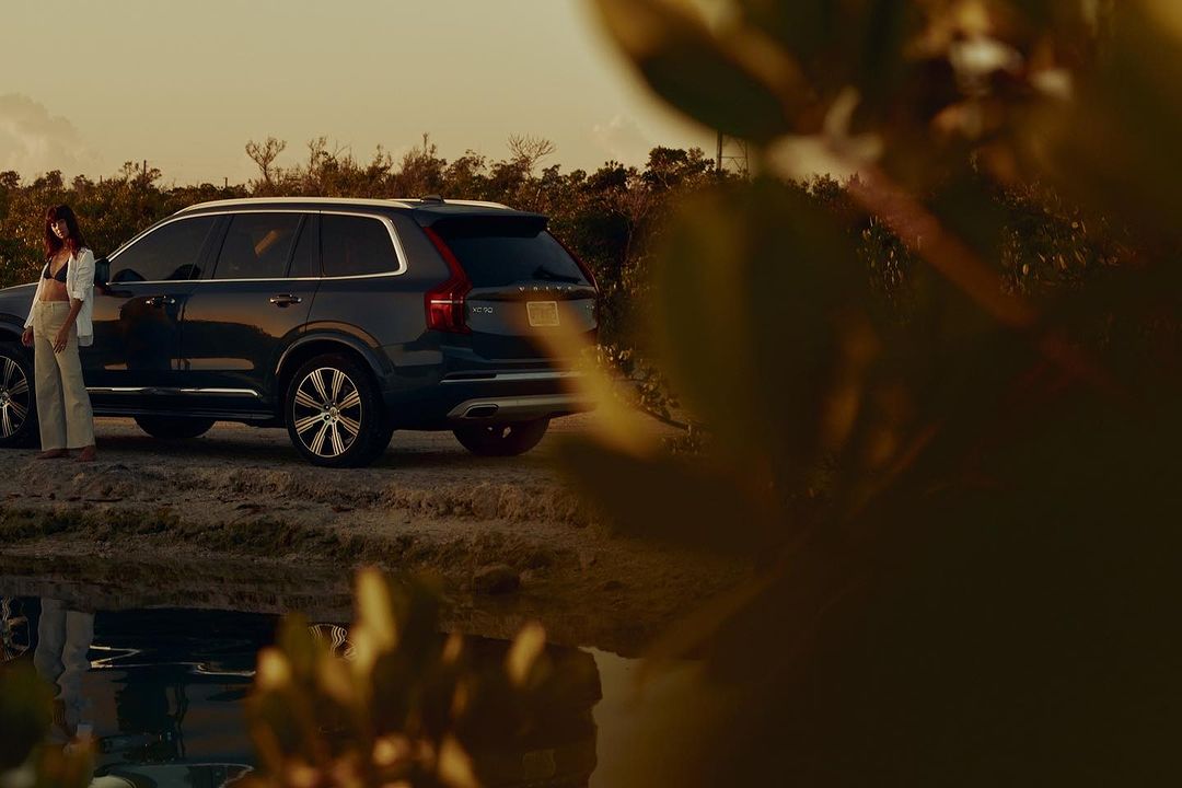 An adult female stands next to a dark blue Volvo SUV on a sandy shore at sunset with vegetation in the background.