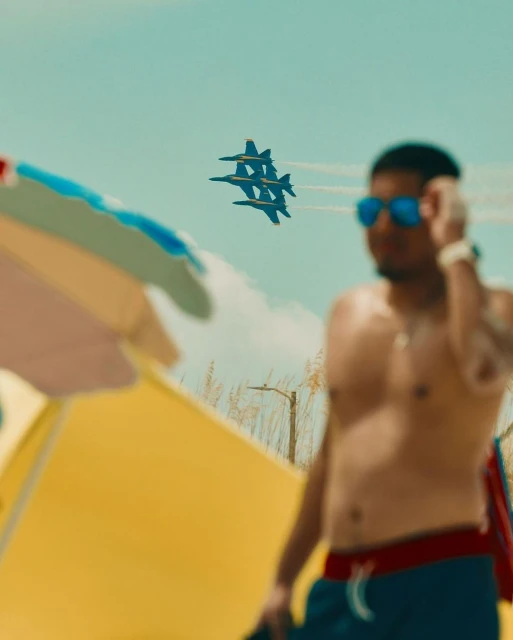 A man with sunglasses stands near the beach with military planes overhead, creating a dynamic scene with a tropical atmosphere.