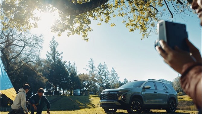 Olive-green SUV is parked in grassy field near two males setting up a blue and yellow tent, while a woman is taking a photo.