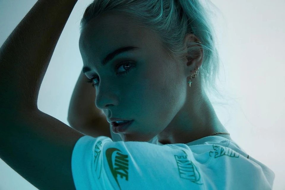 Close-up portrait of a young adult woman with light blue hair wearing a white Nike t-shirt, standing indoors under blue lighting.