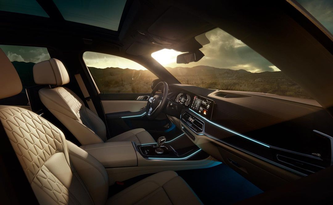 Beige leather interior of a BMW X7 SUV with quilted design, illuminated console, dashboard, and a view of a scenic landscape at sunset.