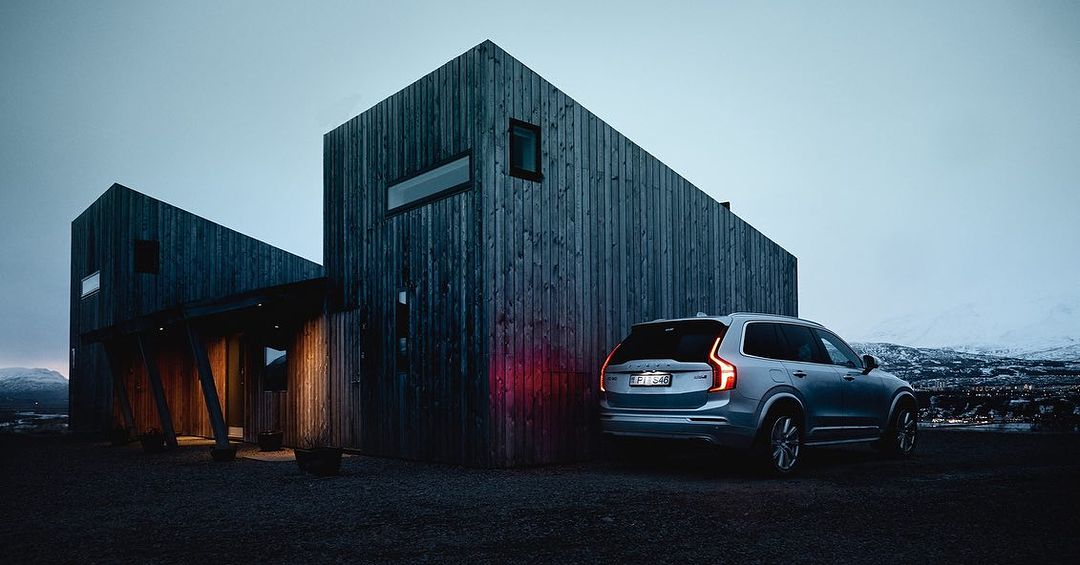 Modern dark wood house and a silver Volvo SUV at dusk with a snowy mountain background, showcasing architectural design and automotive photography.