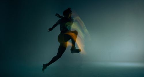 Silhouette of an athletic woman with long exposure motion blur leaping in mid-air against a muted blue gradient background.