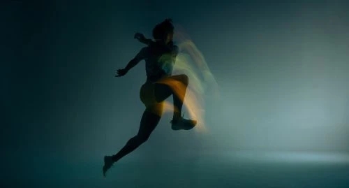 Silhouette of an athletic woman with long exposure motion blur leaping in mid-air against a muted blue gradient background.