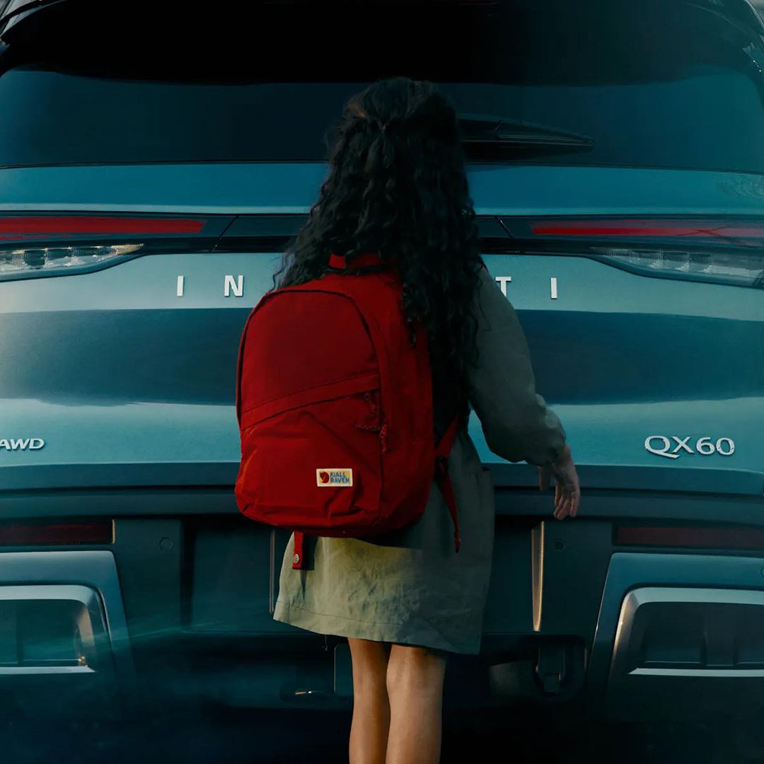 Rear view of an Infiniti QX60 with a young girl wearing a backpack, facing the car, suggesting a travel or family outing.
