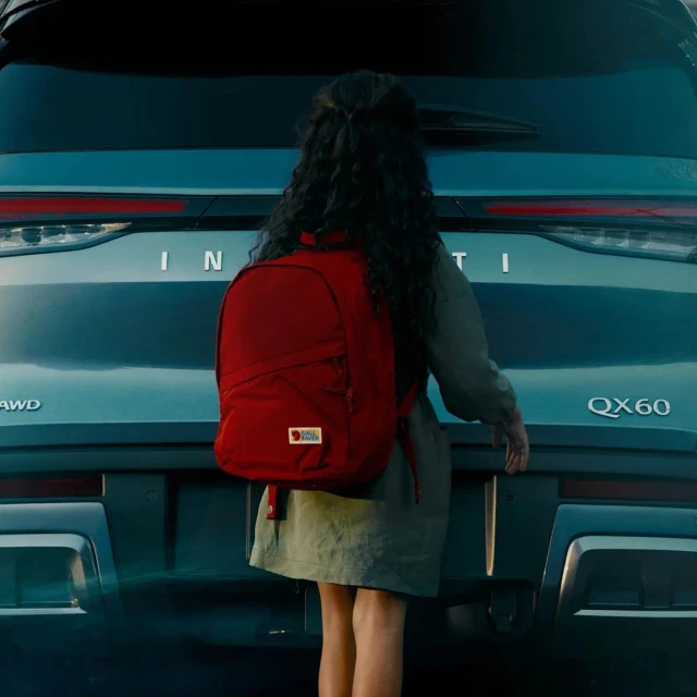 Rear view of an Infiniti QX60 with a young girl wearing a backpack, facing the car, suggesting a travel or family outing.