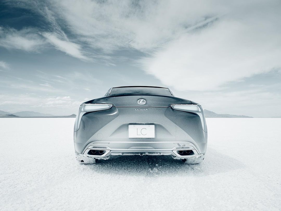 Rear view of a silver Lexus LC 500 coupe on a white salt flat under a cloudy sky. The vehicle's taillights and logo are visible.