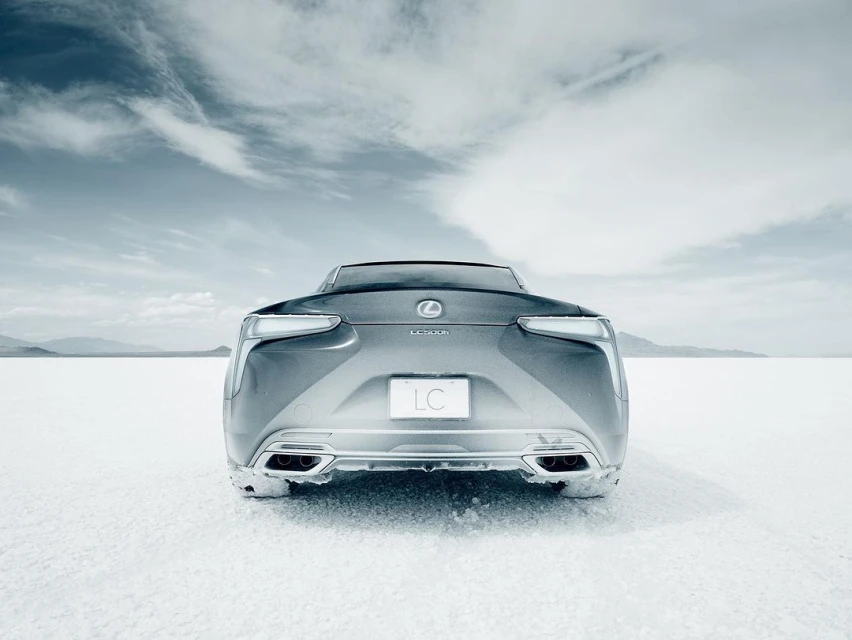 Rear view of a silver Lexus LC 500 coupe on a white salt flat under a cloudy sky. The vehicle's taillights and logo are visible.