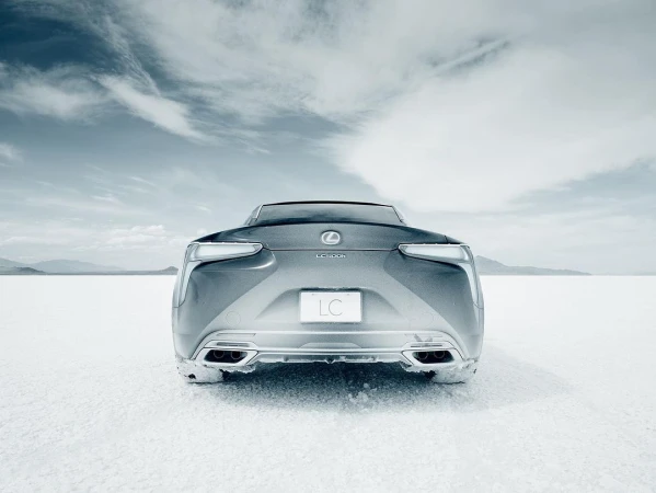 Rear view of a silver Lexus LC 500 coupe on a white salt flat under a cloudy sky. The vehicle's taillights and logo are visible.