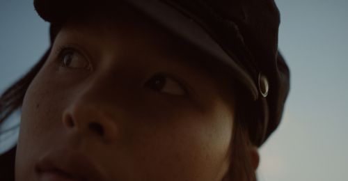 Close-up portrait of a woman wearing a cap, looking to the side against a blue sky backdrop, showcasing a fashion-forward aesthetic.