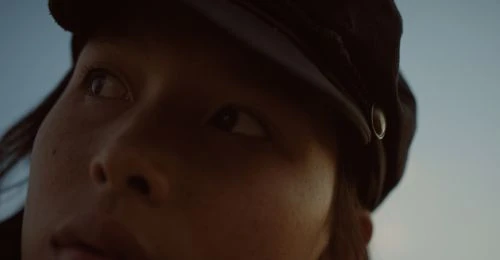 Close-up portrait of a woman wearing a cap, looking to the side against a blue sky backdrop, showcasing a fashion-forward aesthetic.
