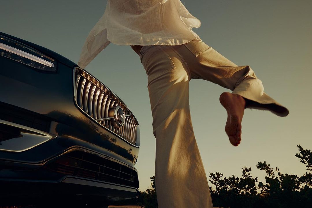 Woman stands on a black Volvo SUV on a beach under a sunny sky, showcasing automotive lifestyle and beachside elegance.