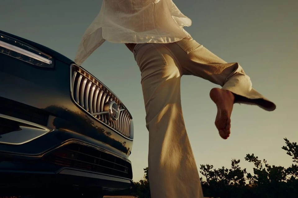 Woman stands on a black Volvo SUV on a beach under a sunny sky, showcasing automotive lifestyle and beachside elegance.