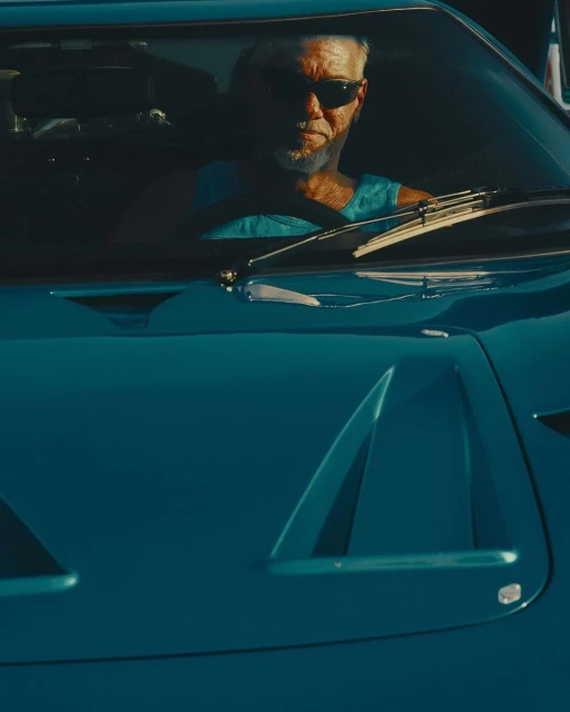 Older man driving a vintage-style blue car with visible hood vents, wearing sunglasses, and a tank top on a sunny day.