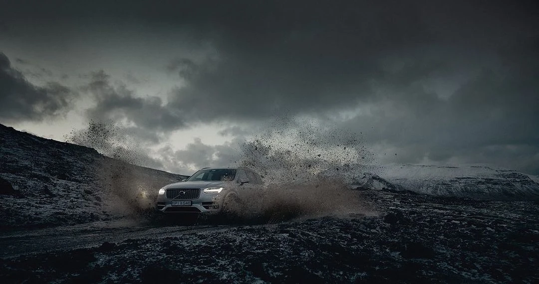 Silver Volvo SUV driving through a mud puddle with water splashing against a backdrop of snowy mountains under a cloudy sky.