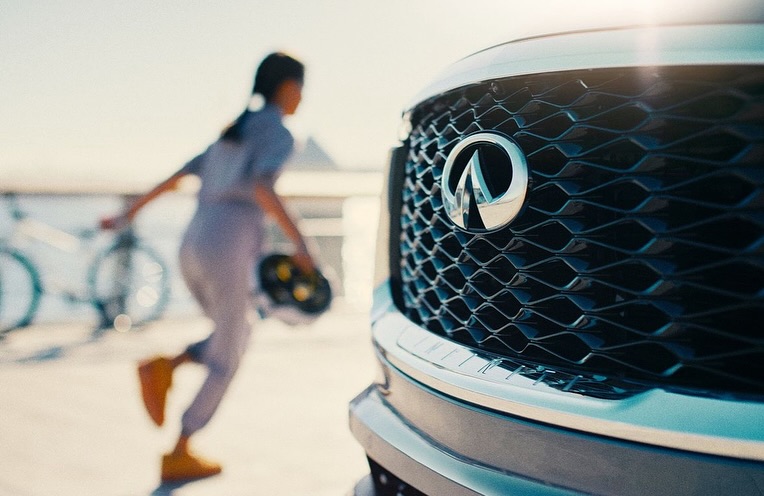 Car's front grille with the Infiniti logo in focus; a woman with a helmet walks by with bicycles blurred in the background.