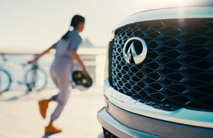 Car's front grille with the Infiniti logo in focus; a woman with a helmet walks by with bicycles blurred in the background.