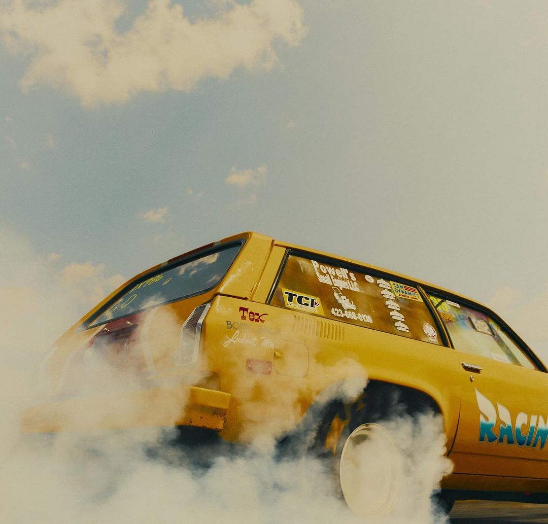 Yellow vintage station wagon car with racing decals performs tire burnout, generating smoke cloud against a blue and cloudy sky outdoors.